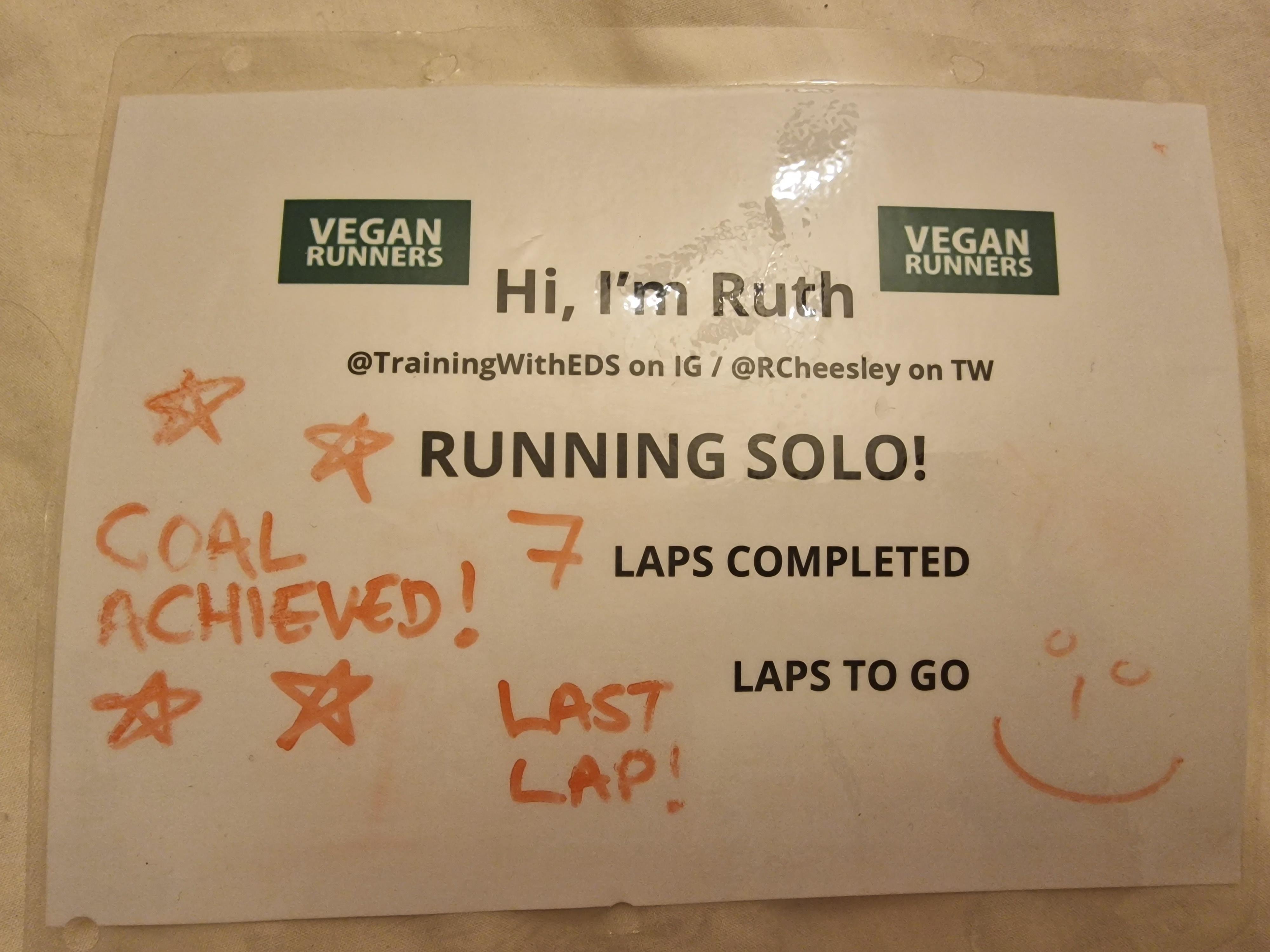 Photo of a laminated sign saying 'Hi I'm Ruth, running solo, 7 laps completed, 0 laps to go with 'Goal achieved' and 'Last lap' written in red pen and vegan runners logos on the top.