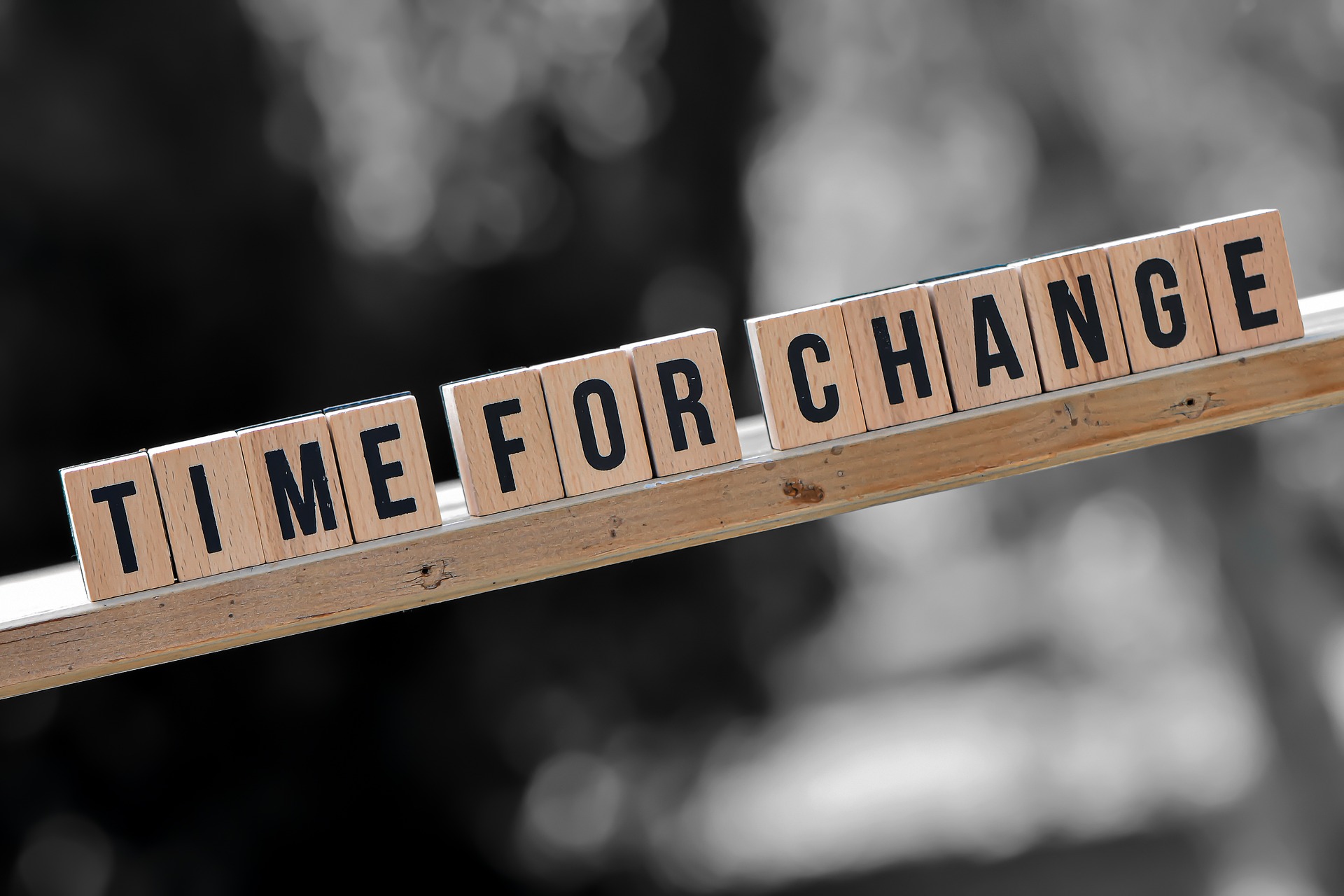 A photo of wooden word tiles spelling out 'time for change' on a wooden surface at an angle, with a blurred black and white background