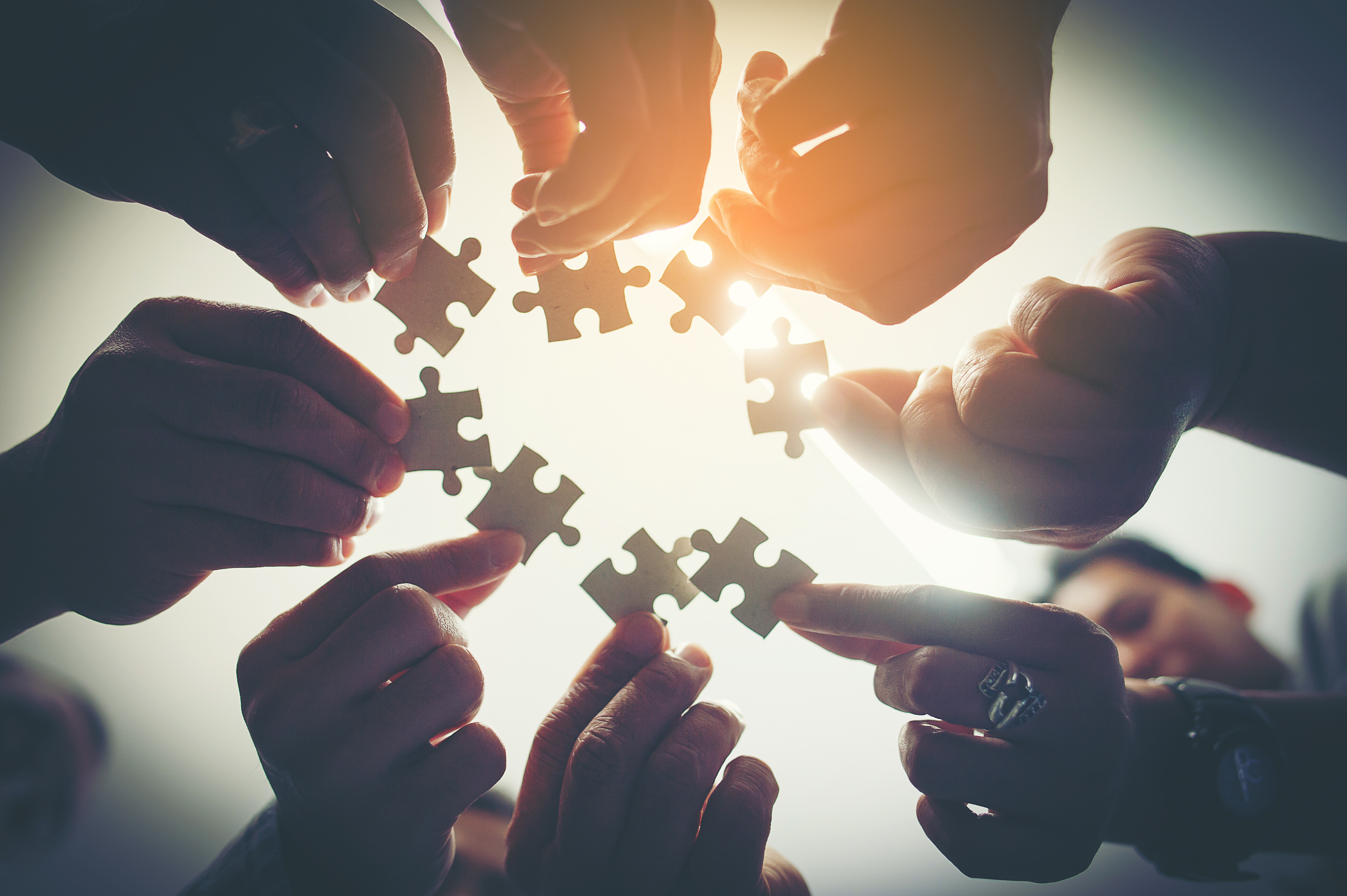 A photo of several people standing in a circle with their hands each holding a piece of a jigsaw with a light source above and behind them