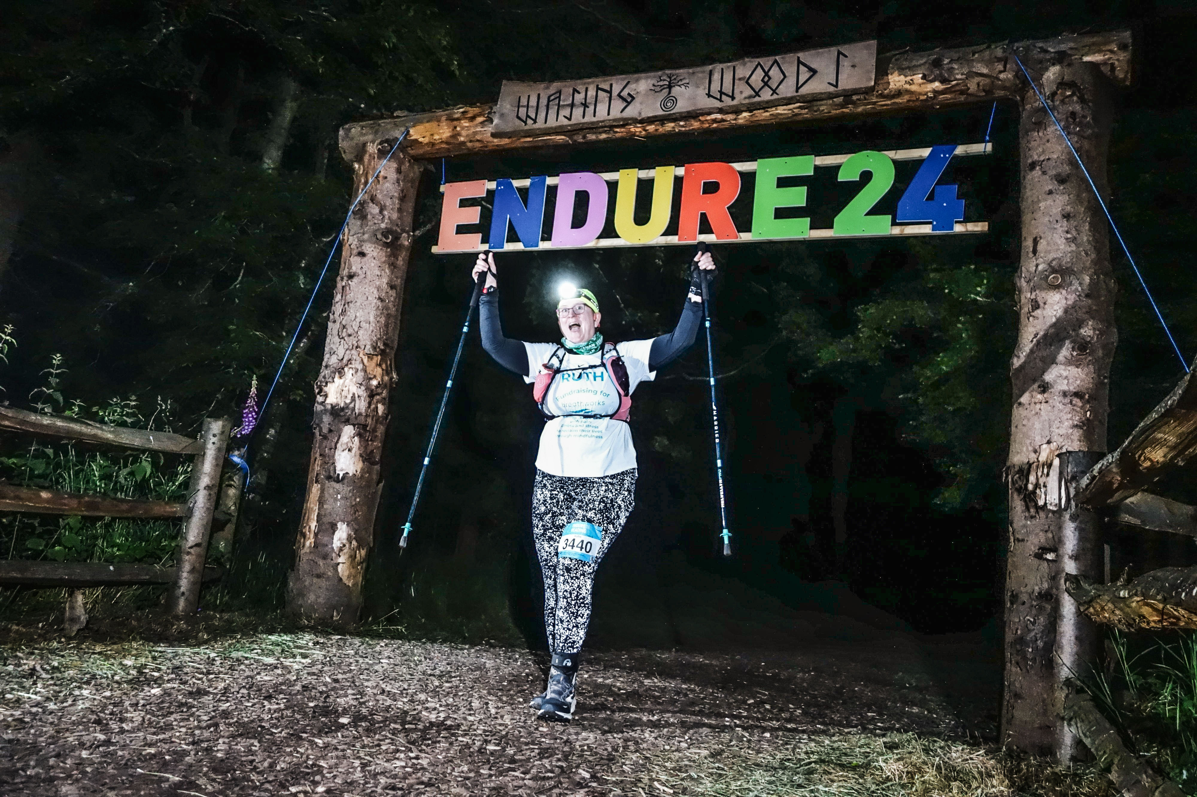Photo of Ruth at night in the dark coming through the Wasing Woods gate. She's got a headtorch on her head which is switched on, and reflective leggings which are showing bright white dots. She's holding both her poles up in the air in an expression of delight.