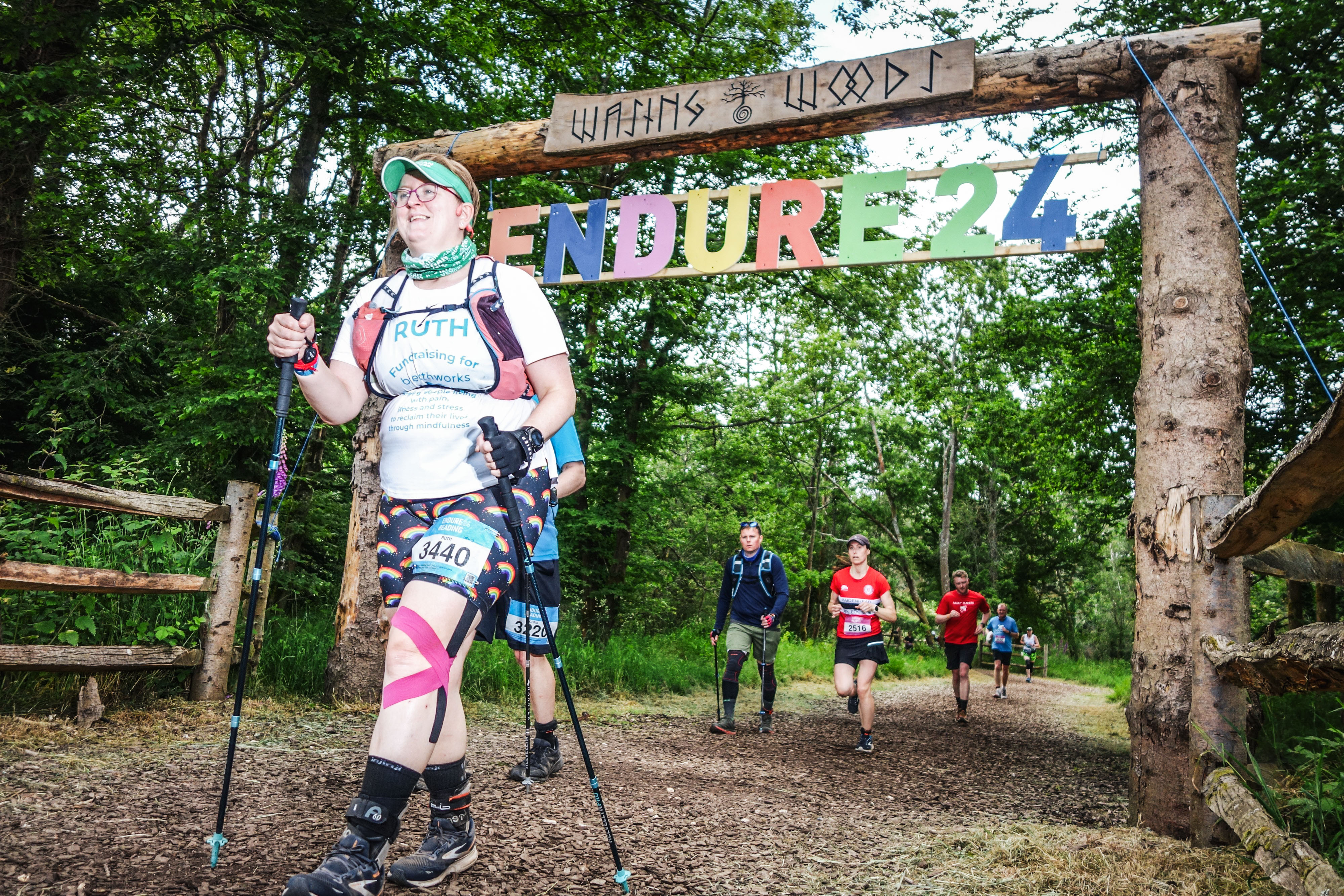 Photo of Ruth coming through the gate at Wasing Woods, she's smiling and holding two walking poles with a heavily taped left knee.