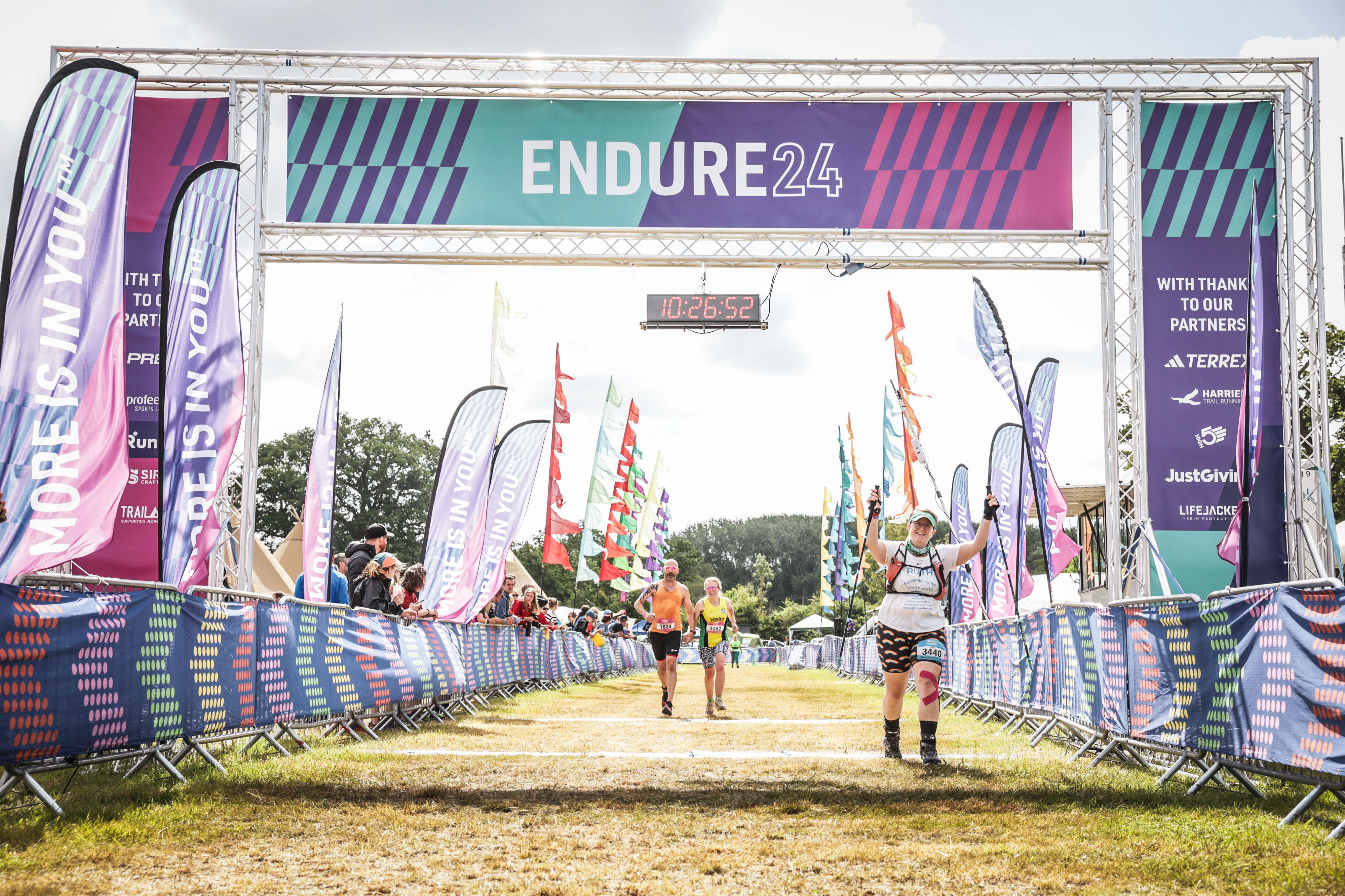 Photo of Ruth coming over the finish line for the last time, the clock shows the time of 10:26:52 and she's holding both hands up in triumph and smiling at the camera.