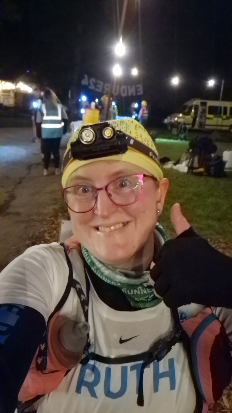 Ruth at night with lots of bright lights behind her at an aid station, she's smiling at the camera with one thumb raised.