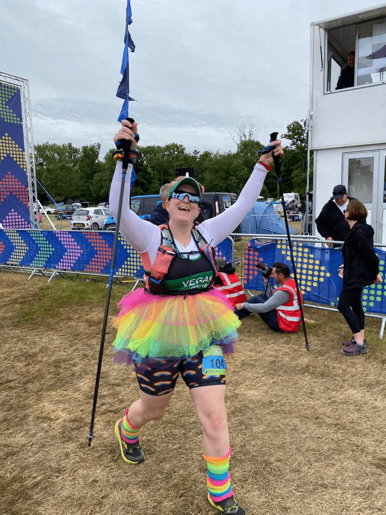 Ruth finishing Endure24 wearing a black and green Vegan Runners vest, a luminous rainbow tutu with a fluffy luminous tail attached, luminous rainbow leg warmers and a unicorn hairband. She has her arms in the air and she is smiling.