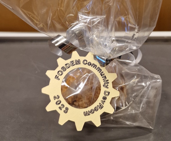 Photo of cake in a cellophane wrapper with a cut out cog shaped label with 'FOSDEM Community Dev Room 2023' on the edge of the cog.