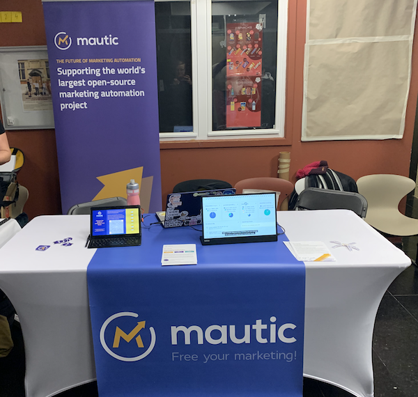 A photo of the Mautic stand at FOSDEM. There's a blue runner over a white table with the Mautic logo on it, and a popup banner standing behind the table. On the table is a tablet with a landing page and QR code, and a monitor which is showing a video of Mautic.