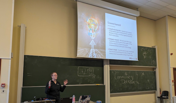 A photo of Ruth at the front of the auditorium in mid-flow delivering her talk. On the screen behind and to the right is a slide explaining the lessons learned in a part of the process of changing the governance model.