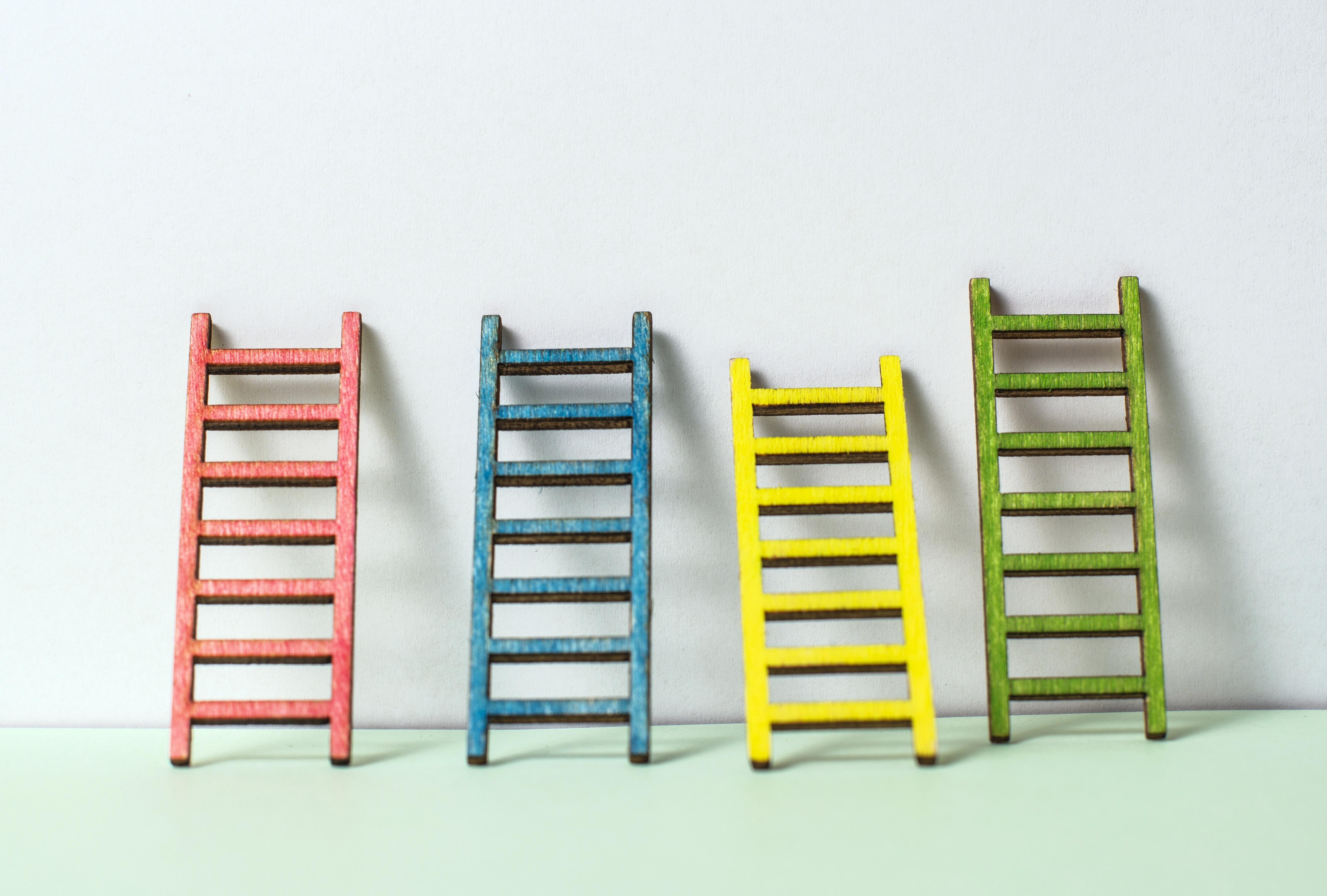 Photo of four ladders in red, blue, yellow and green leaning up against a white wall on a pale green floor.
