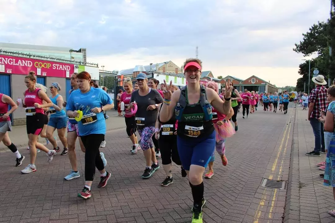 Ruth running in the Ipswich Twilight 10k race wearing a green and black Vegan Runners vest, blue cycling shorts and a pink visor. She's smiling at the camera and making V signs with both hands, surrounded by other runners.