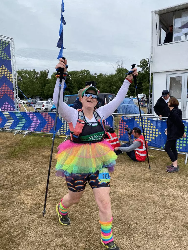 Ruth crossing the finish line at Endure24 with her hands raised in the air, dressed as a rainbow unicorn with a tutu, tail and headband with luminous rainbow leg warmers.