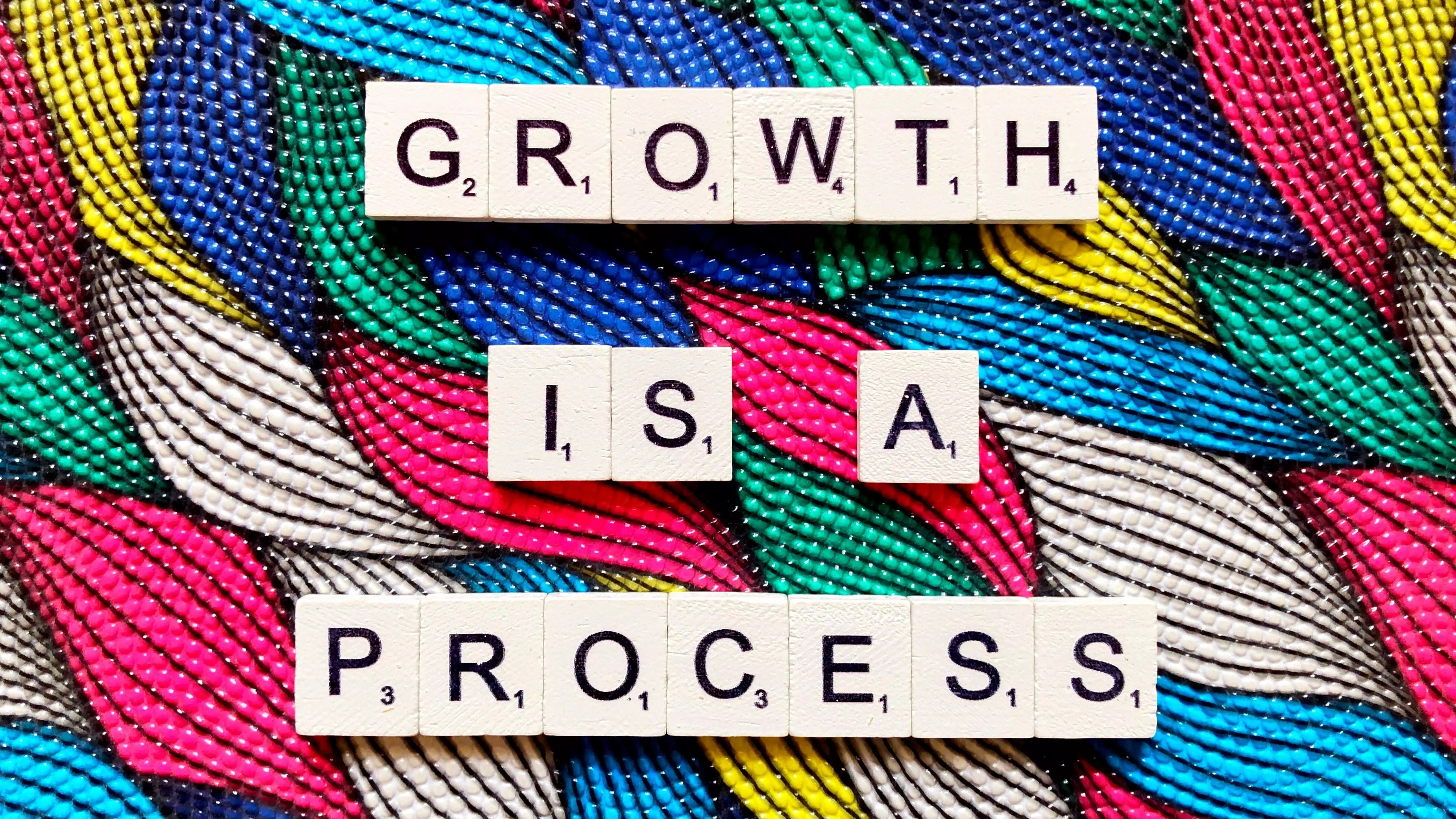 Photo of several brightly coloured woven fabric that are woven into a plait-like pattern, with the words in scrabble letters 'growth is a process' on top.