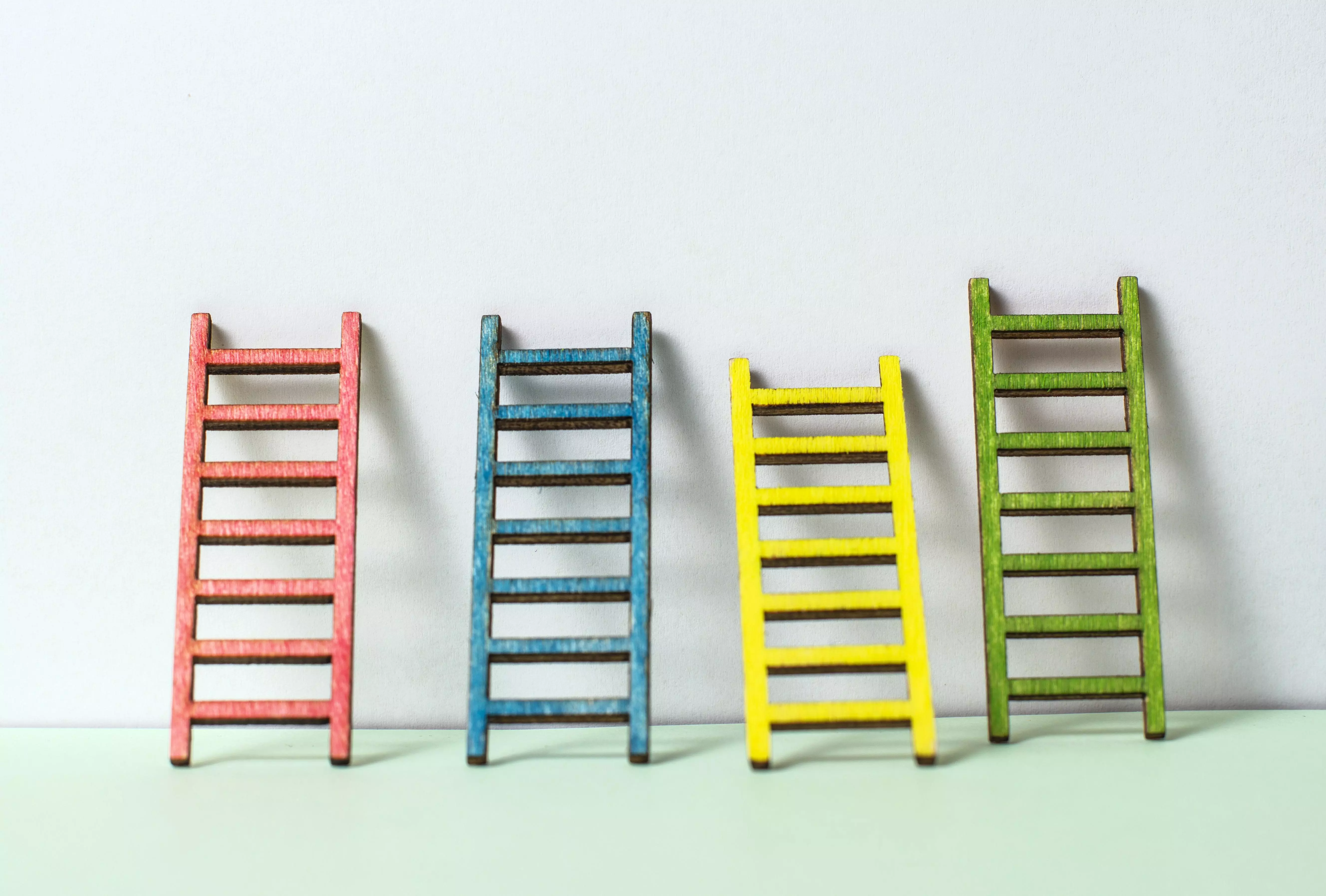 Photo of four ladders in red, blue, yellow and green leaning up against a white wall on a pale green floor.