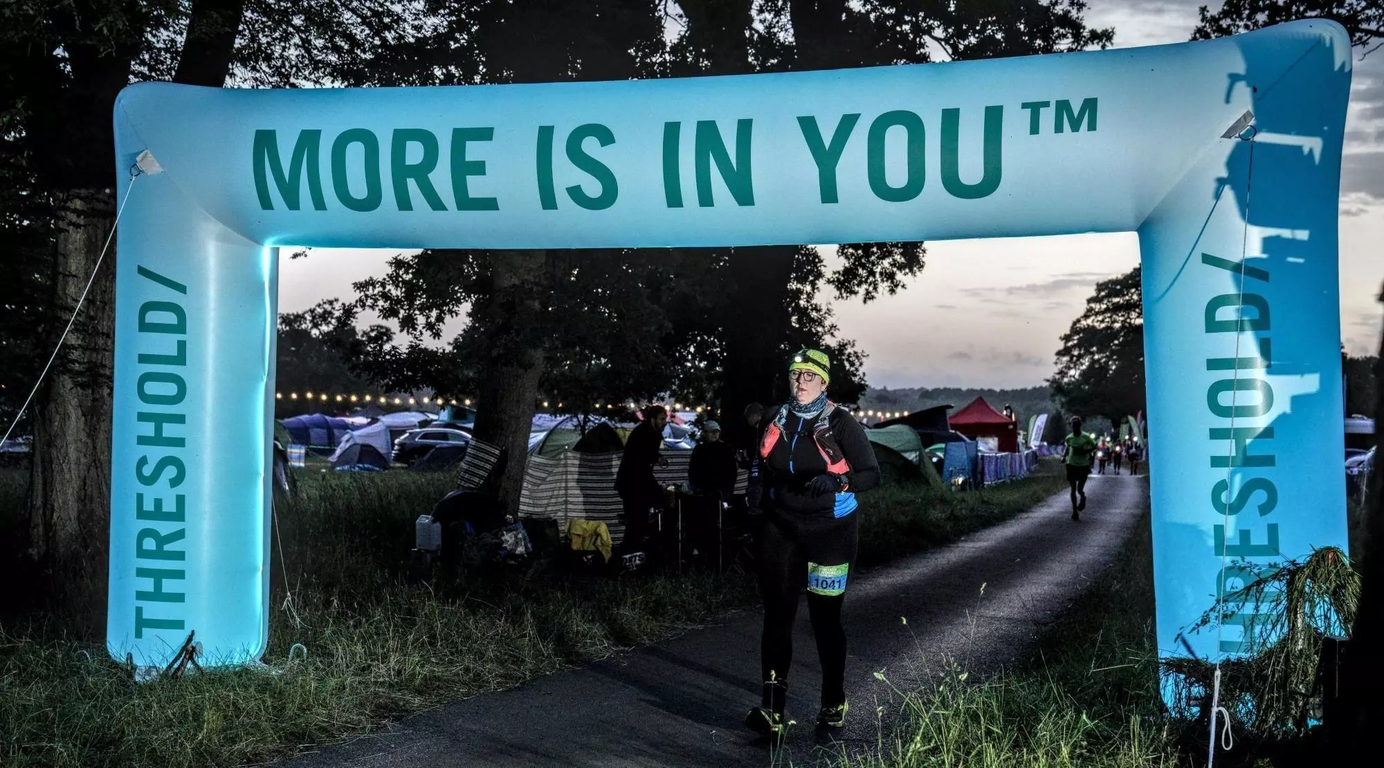Photo of Ruth running at dawn with a headtorch under an illuminated arch saying 'More is in you'.