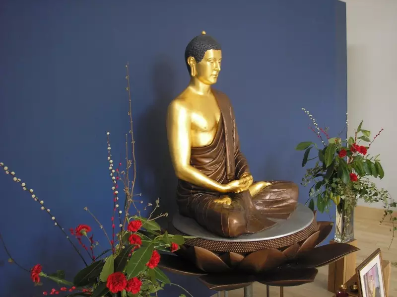 A photo of the Buddha rupa at Ipswich Buddhist Centre, a large golden buddha with hands in meditation posture, with a deep blue backdrop and flowers around the shrine area.