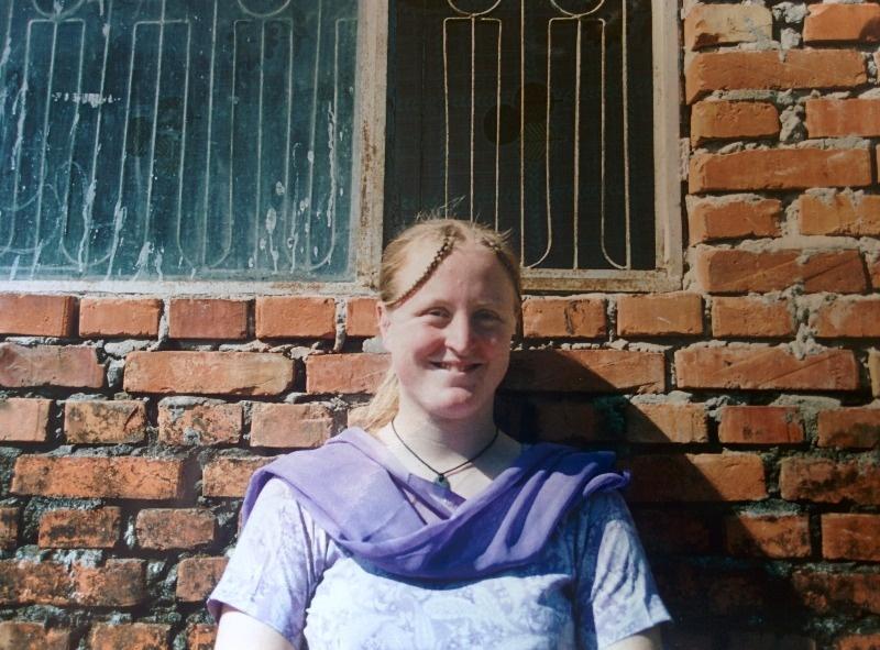Photo of Ruth in front of a roughly built brick wall. She is wearing a purple paisly print salwaar kameez with a purple scarf and has two plaits at the front of long blonde hair which is tied back in a ponytail. She is smiling at the camera.
