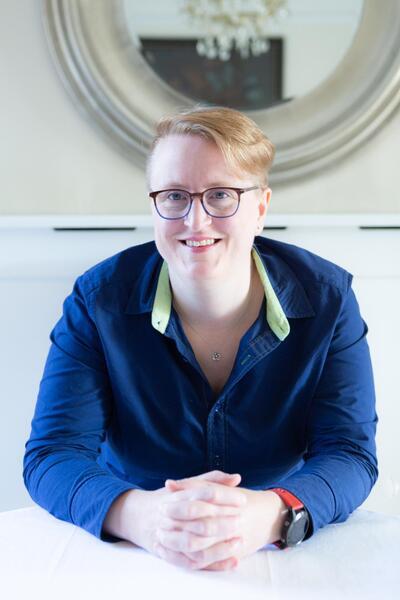 Photo of Ruth facing the camera with her hands in front, leaning slightly forward and smiling. She wears a dark blue long sleeved shirt with a contrasting green inner collar.